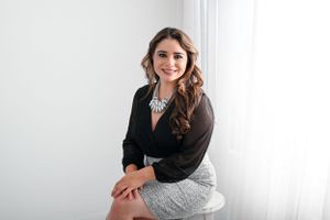 Professional portrait of someone in a black blazer and patterned skirt sitting against a white wall background.