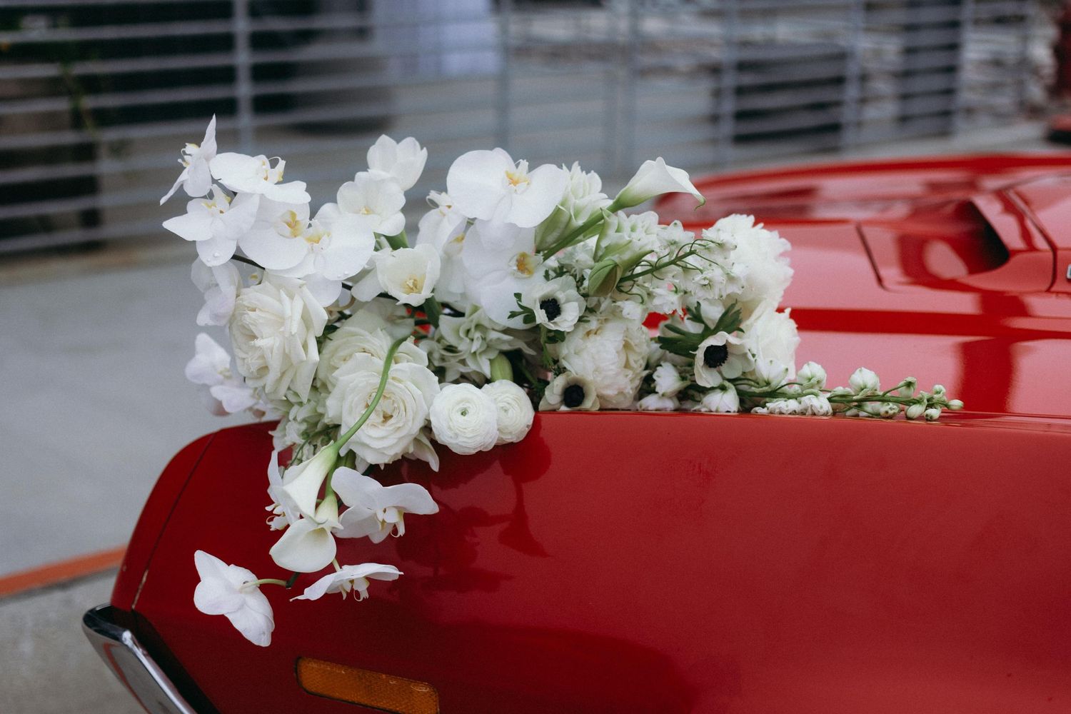 A delicate white flower arrangement rests on the glossy red hood of a car in soft natural lighting.