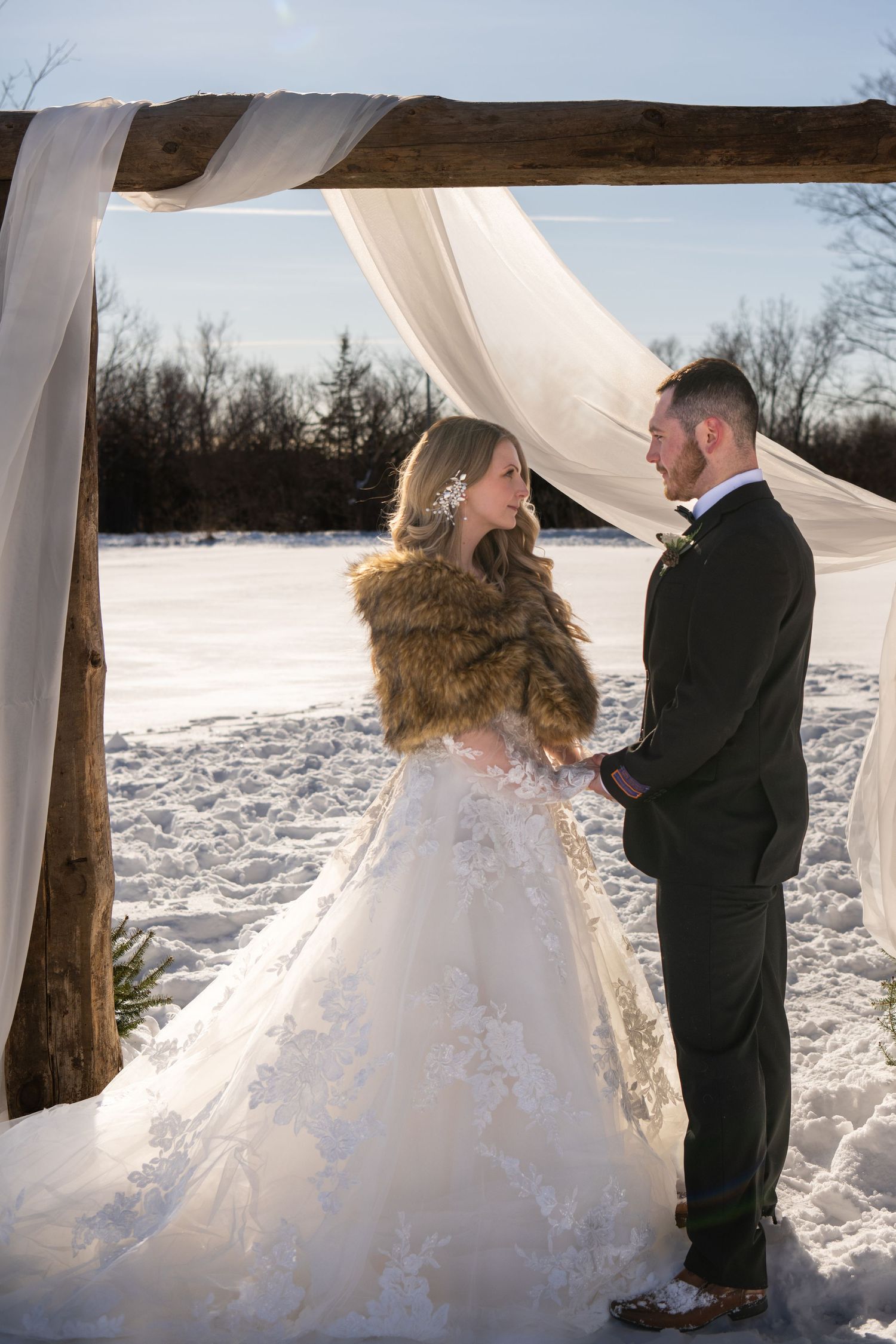 A winter wedding scene with a couple under a white draped arch in the snow, the bride wearing a fur shawl and lace gown.