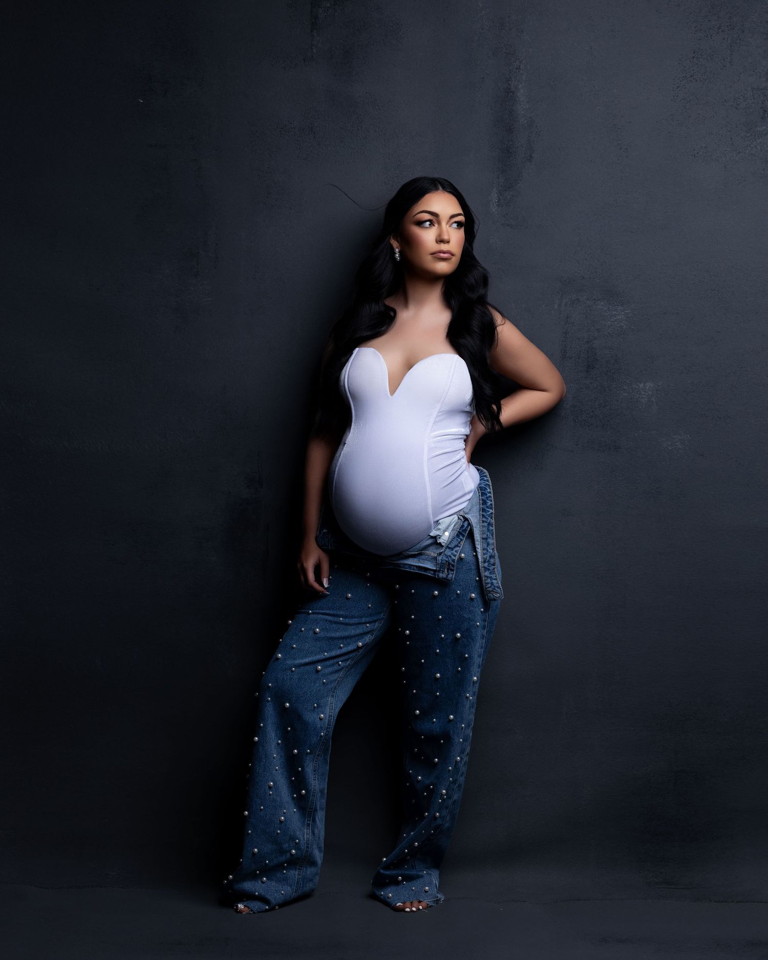 Maternity portrait against dark background showing pregnant figure in white bodysuit and embellished jeans.