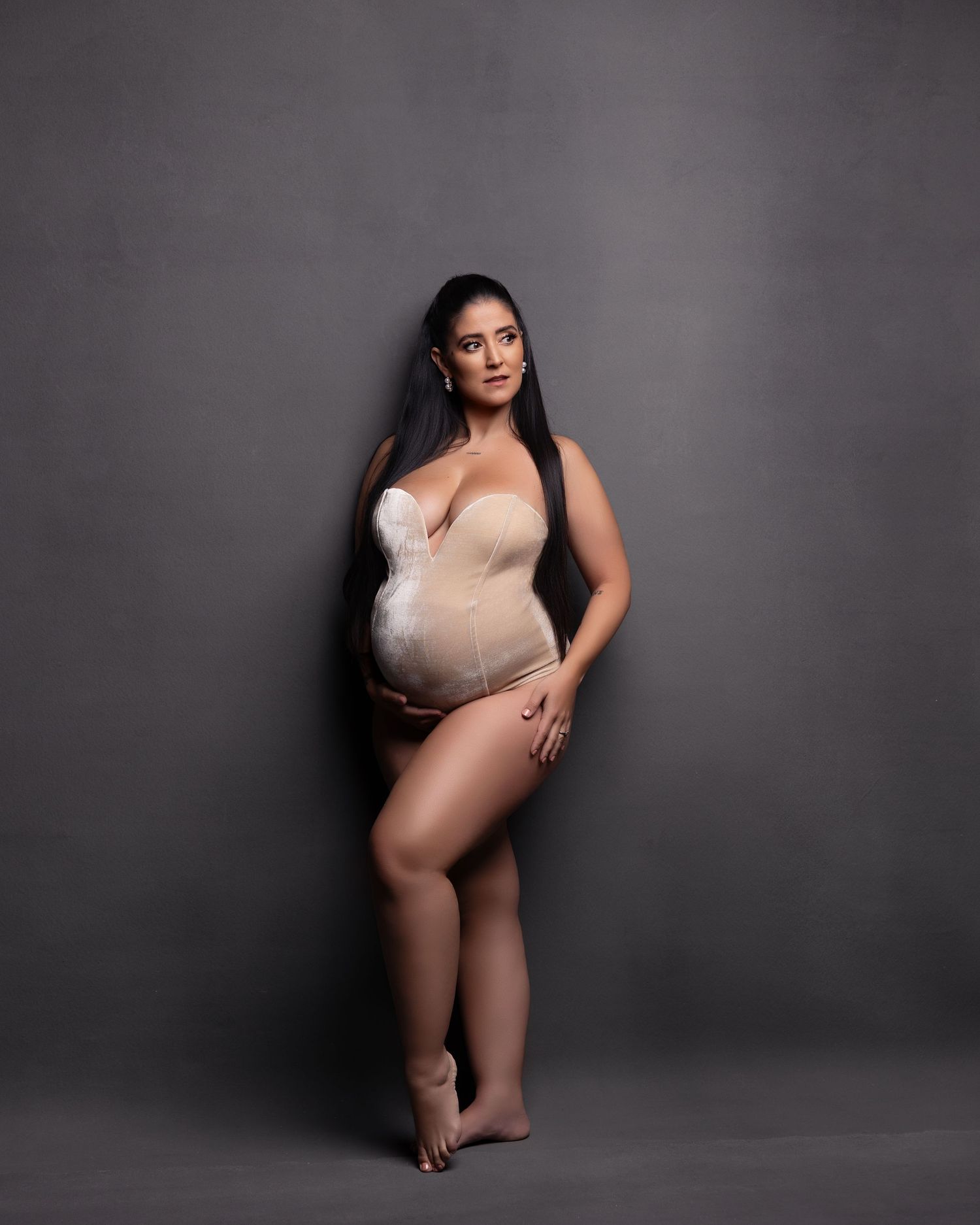 A maternity photo of someone posing in a white bodysuit against a dark gray background in a studio setting.