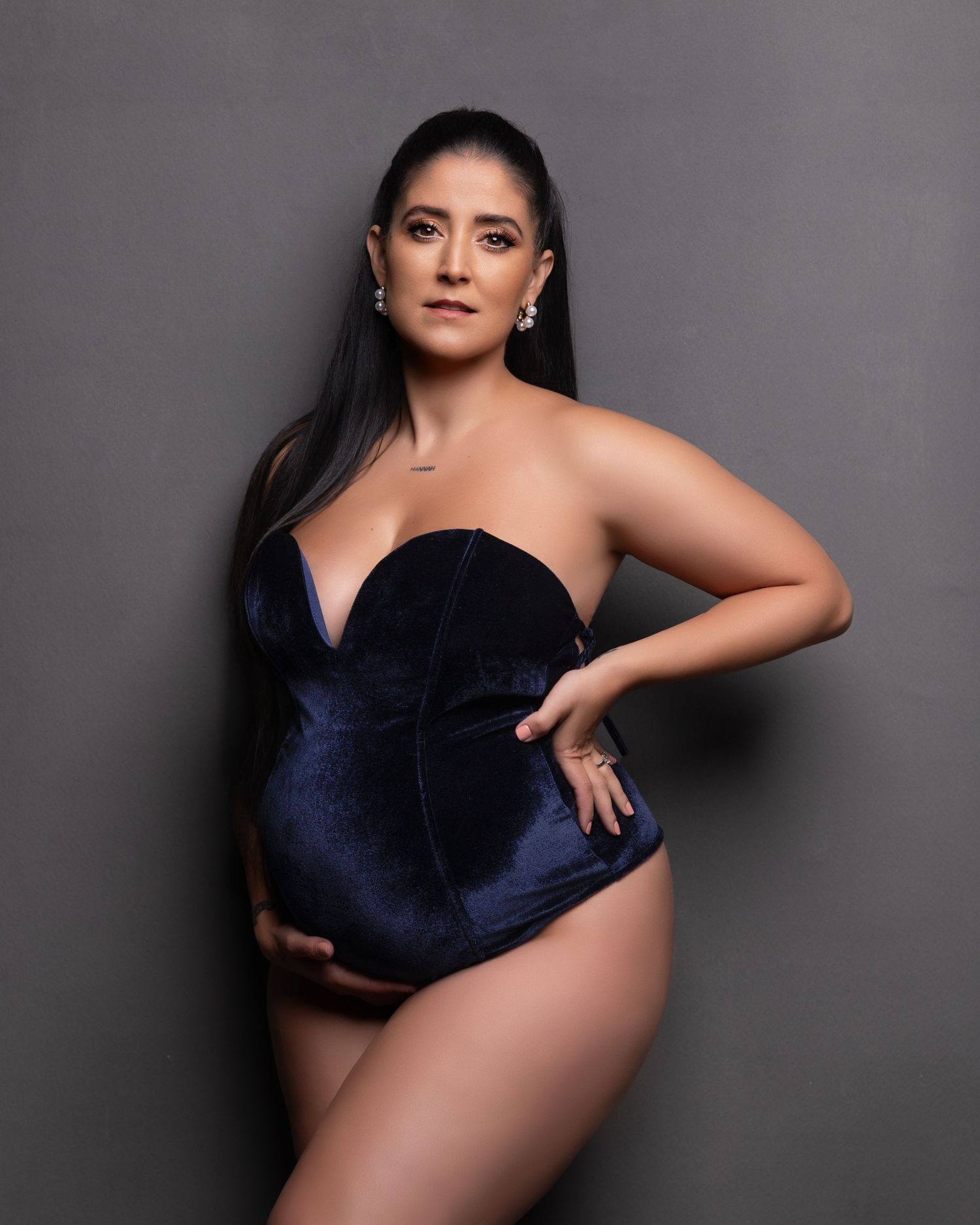 A woman poses in a dark blue velvet bodysuit against a gray studio backdrop for a glamour photoshoot.