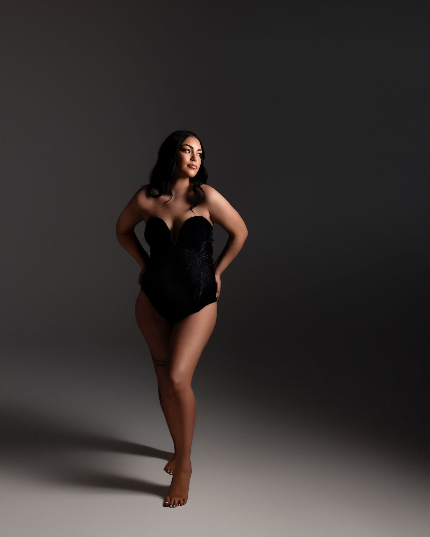 A graceful figure poses in a black corset bodysuit against a dark background with dramatic studio lighting creating shadows.
