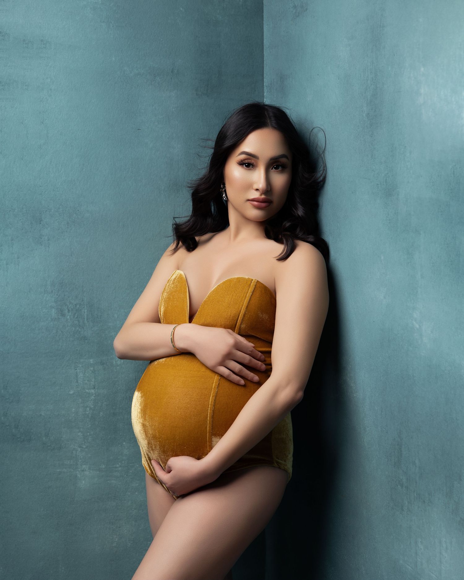 A maternity portrait of a woman in a mustard yellow bodysuit posing against a teal wall in dramatic lighting.