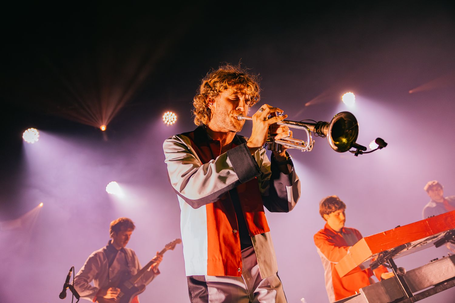 A trumpet player performs with a band on stage under dramatic purple and pink concert lighting.