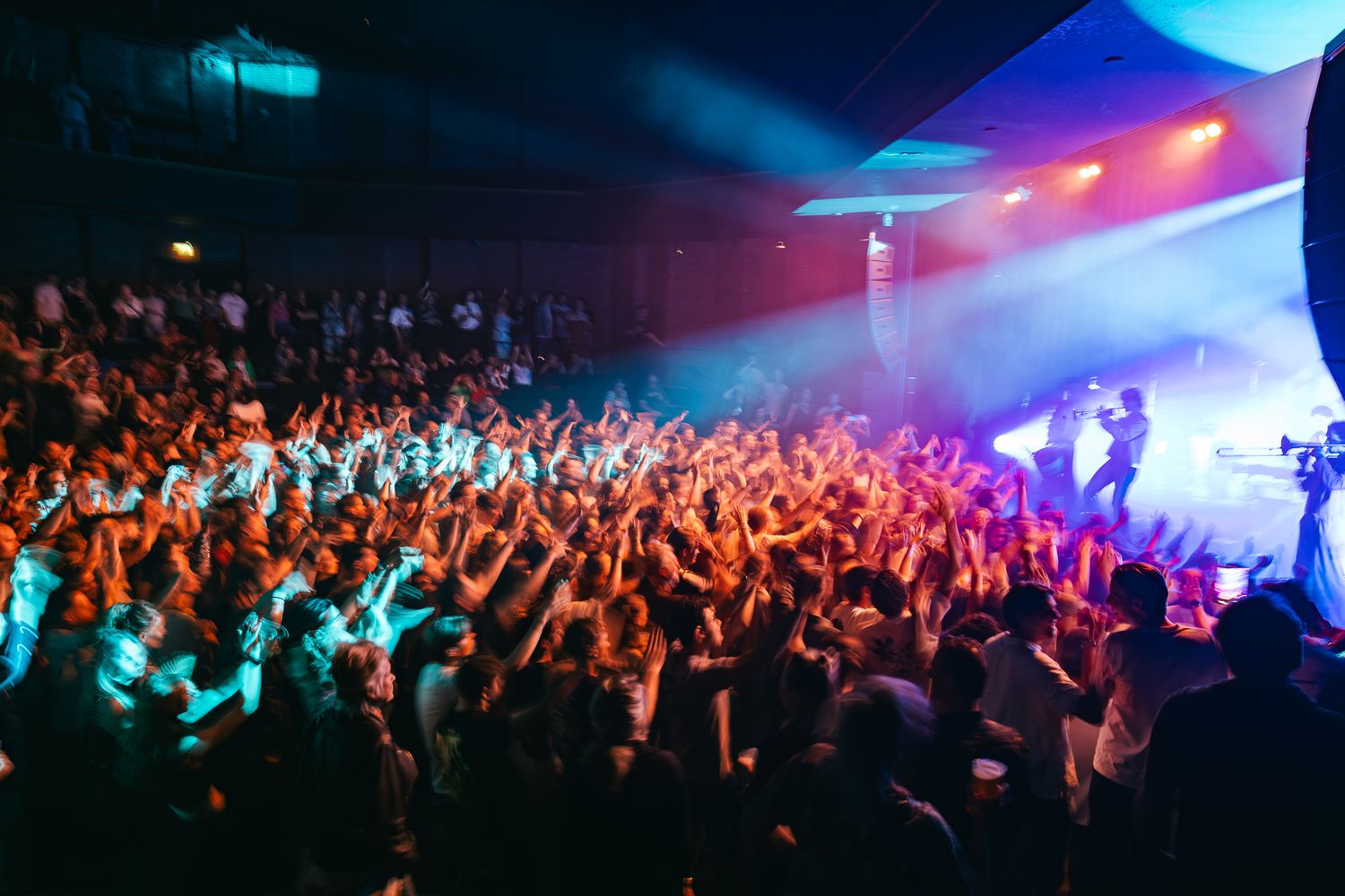 Energetic concert crowd with raised arms under colorful blue and red stage lighting during a live performance.