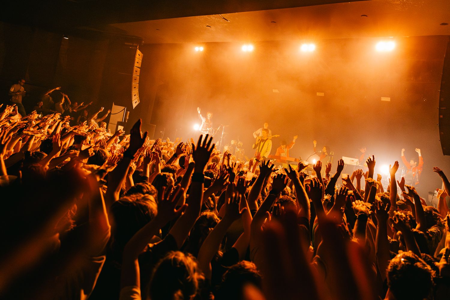 Crowd with raised hands at a concert venue silhouetted against orange stage lighting and fog.