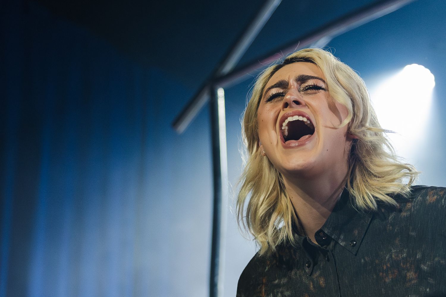 Blonde woman singing passionately on stage under bright spotlight in dark venue.