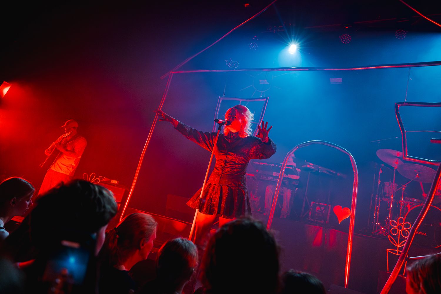 A performer on stage in red and blue lighting at a live music venue, arms outstretched before an energetic crowd.