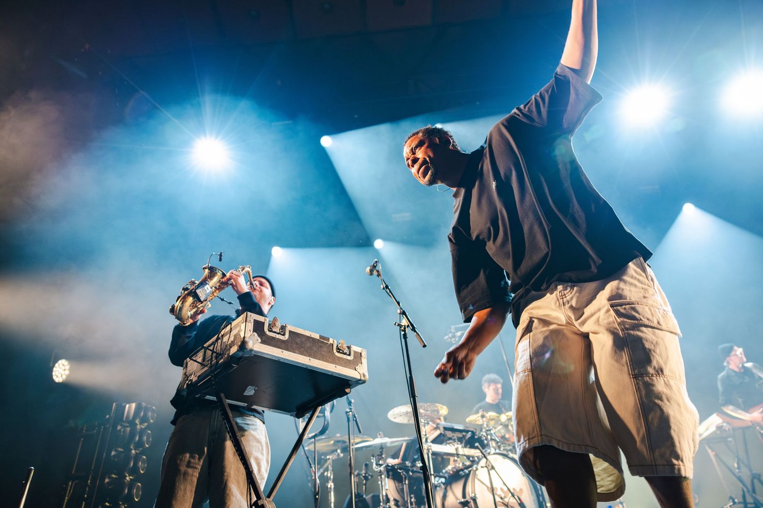 Musicians perform energetically on stage with keyboard and microphone amid dramatic blue lighting and fog effects.