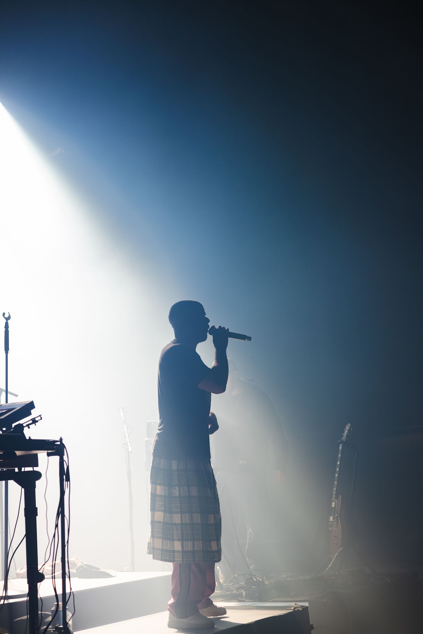 Silhouetted performer in traditional kilt stands on stage with dramatic backlighting and fog effects during concert performance.