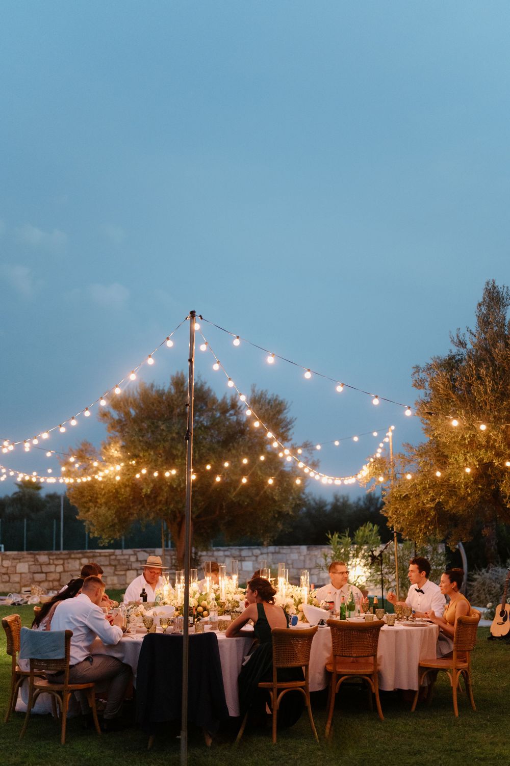 People dining at an outdoor garden party under string lights during dusk with warm ambient lighting and autumn trees.