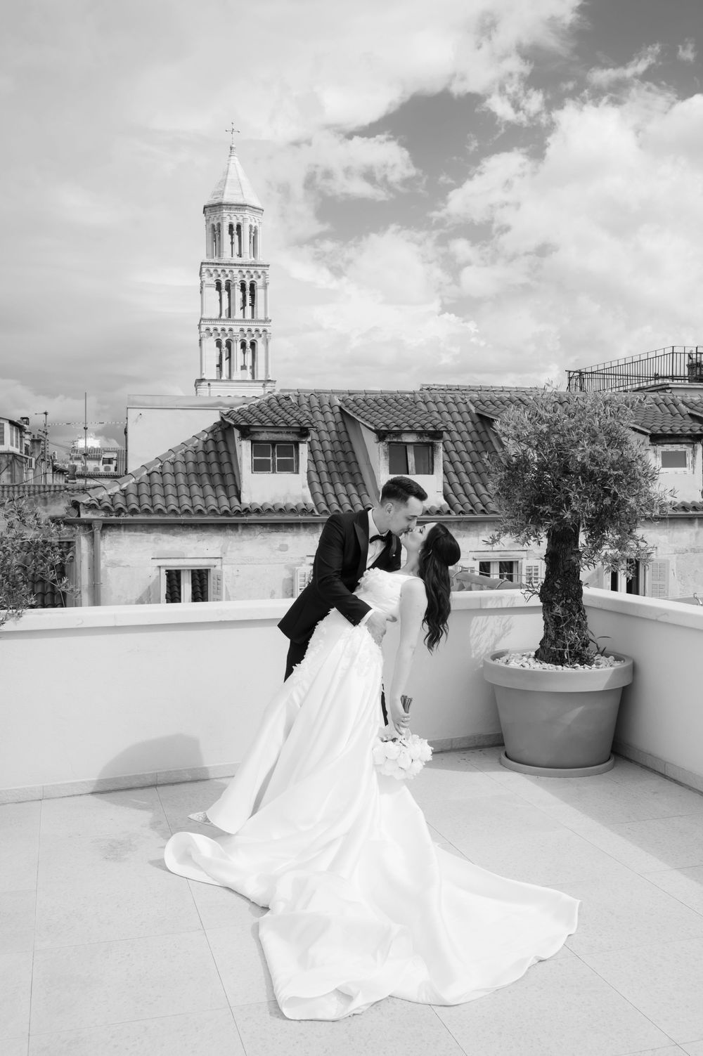 A wedding couple embraces on a rooftop terrace with a historic bell tower and tiled roofs visible in Split, Croatia.