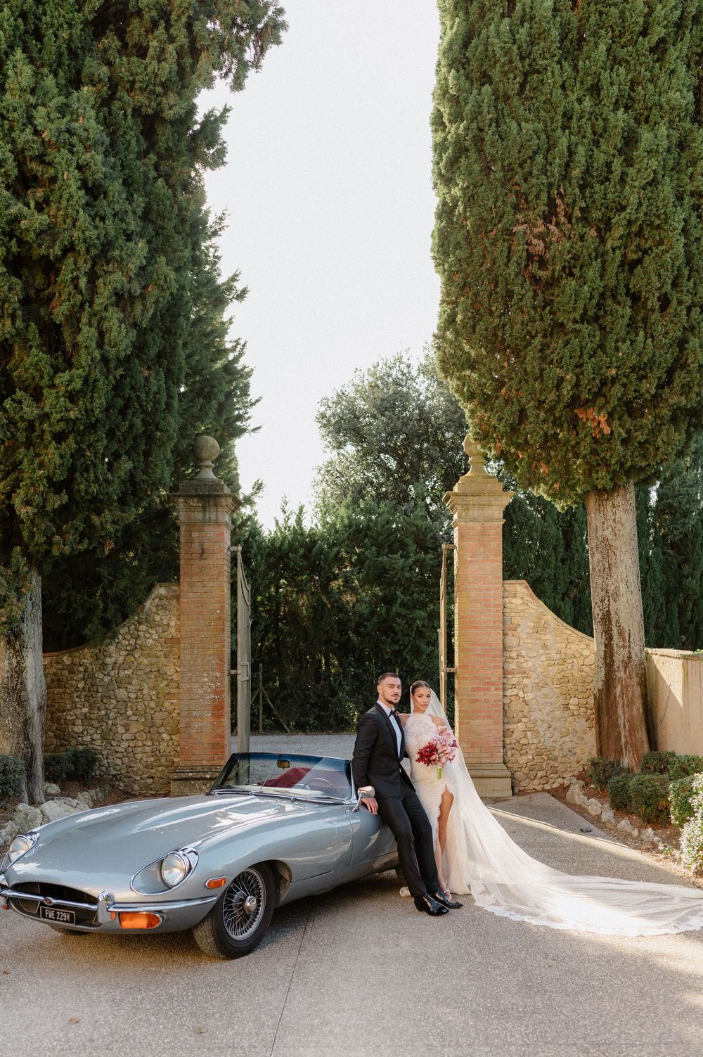 A classic silver Jaguar E-Type parked between stone pillars and cypress trees at an elegant estate entrance.