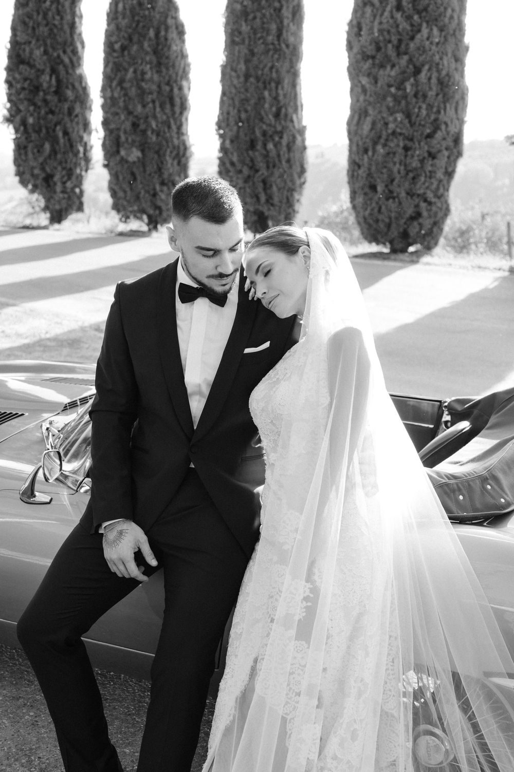 A romantic black and white wedding portrait with cypress trees casting dramatic shadows in the background.