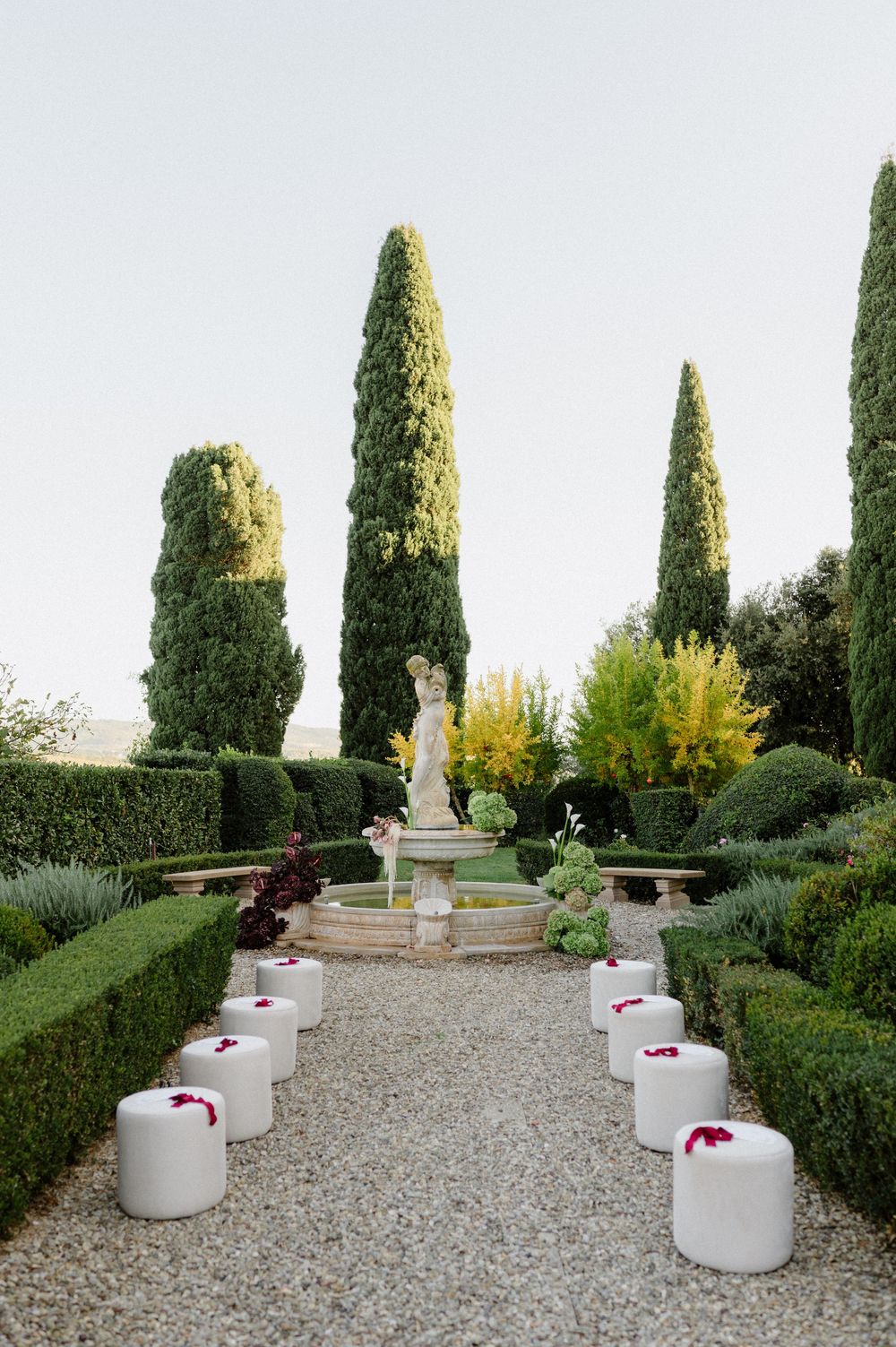 A formal garden path lined with white ceramic stools leads to a classical statue surrounded by tall cypress trees and hedges.