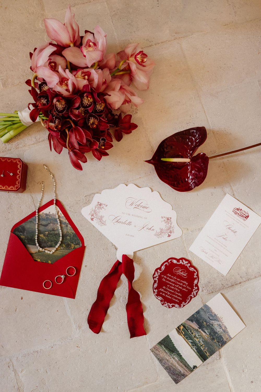 Wedding details flatlay with pink and red flowers, burgundy anthurium, red envelope, rings, and invitation cards on beige surface.