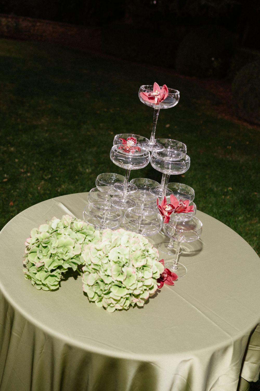 Tiered glass champagne tower being built on round table with green hydrangeas and pink flower accents.
