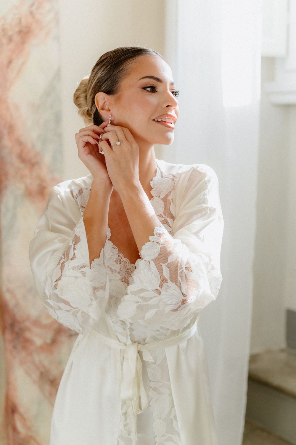 Elegant bride in white lace robe touches her neck while posing by window in soft natural light.