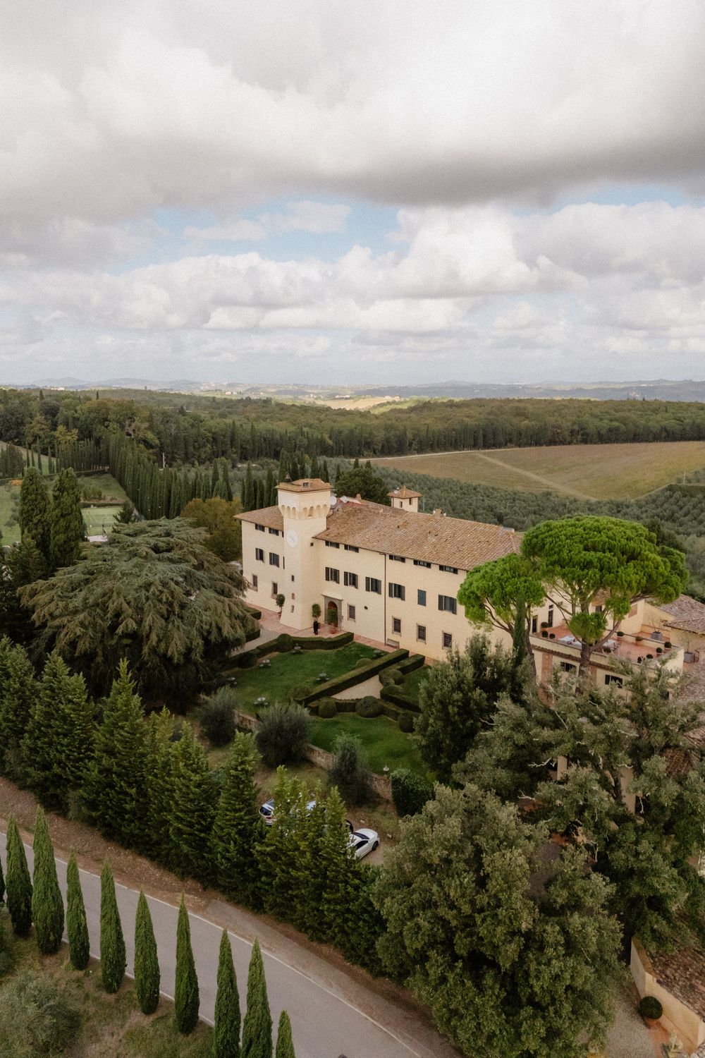 Aerial view of a Tuscan villa nestled in rolling hills with cypress trees lining the driveway under cloudy skies.