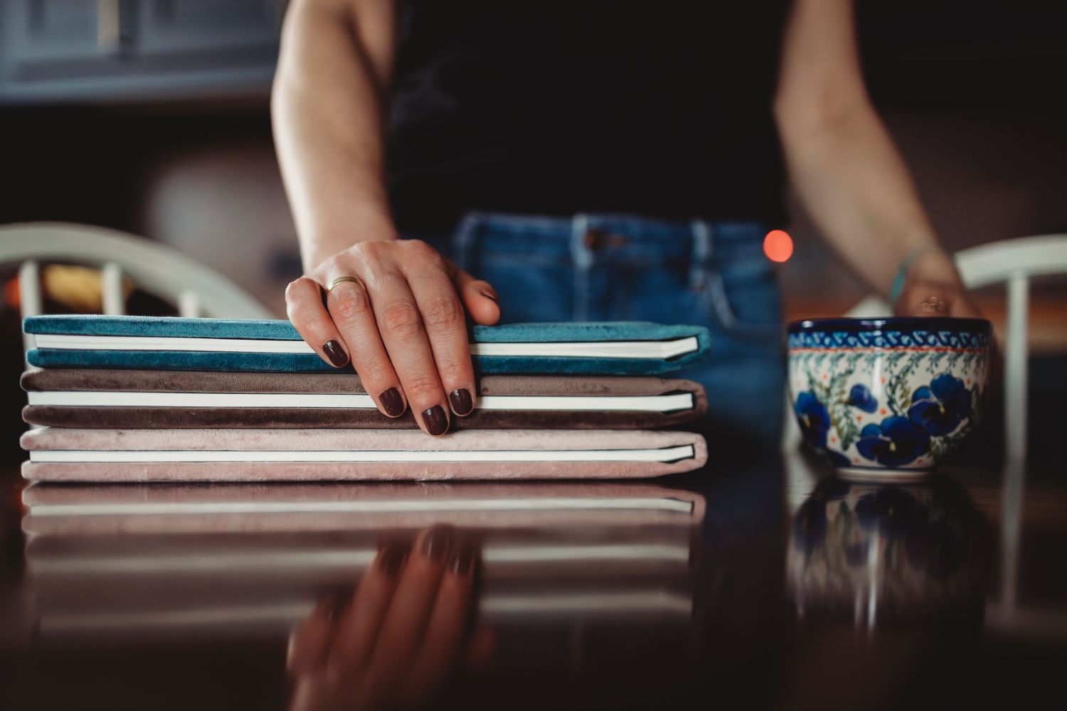 Hand with dark nail polish rests on stack of notebooks beside Earth-themed coffee mug on reflective surface.