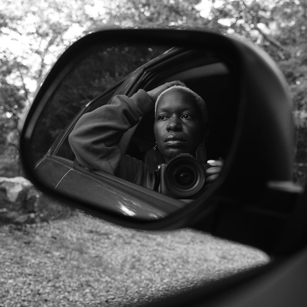 Black and white self-portrait photograph reflected in a car side mirror showing a person holding a camera.