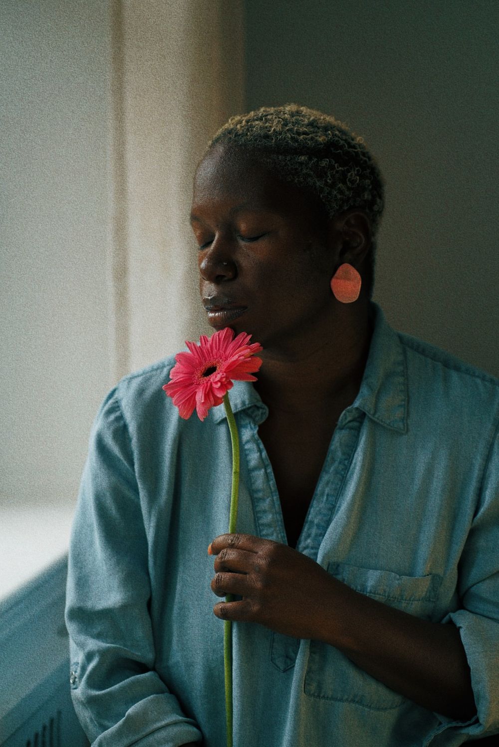 Person wearing turquoise shirt holds single pink gerbera daisy flower while looking to the side in soft window light.
