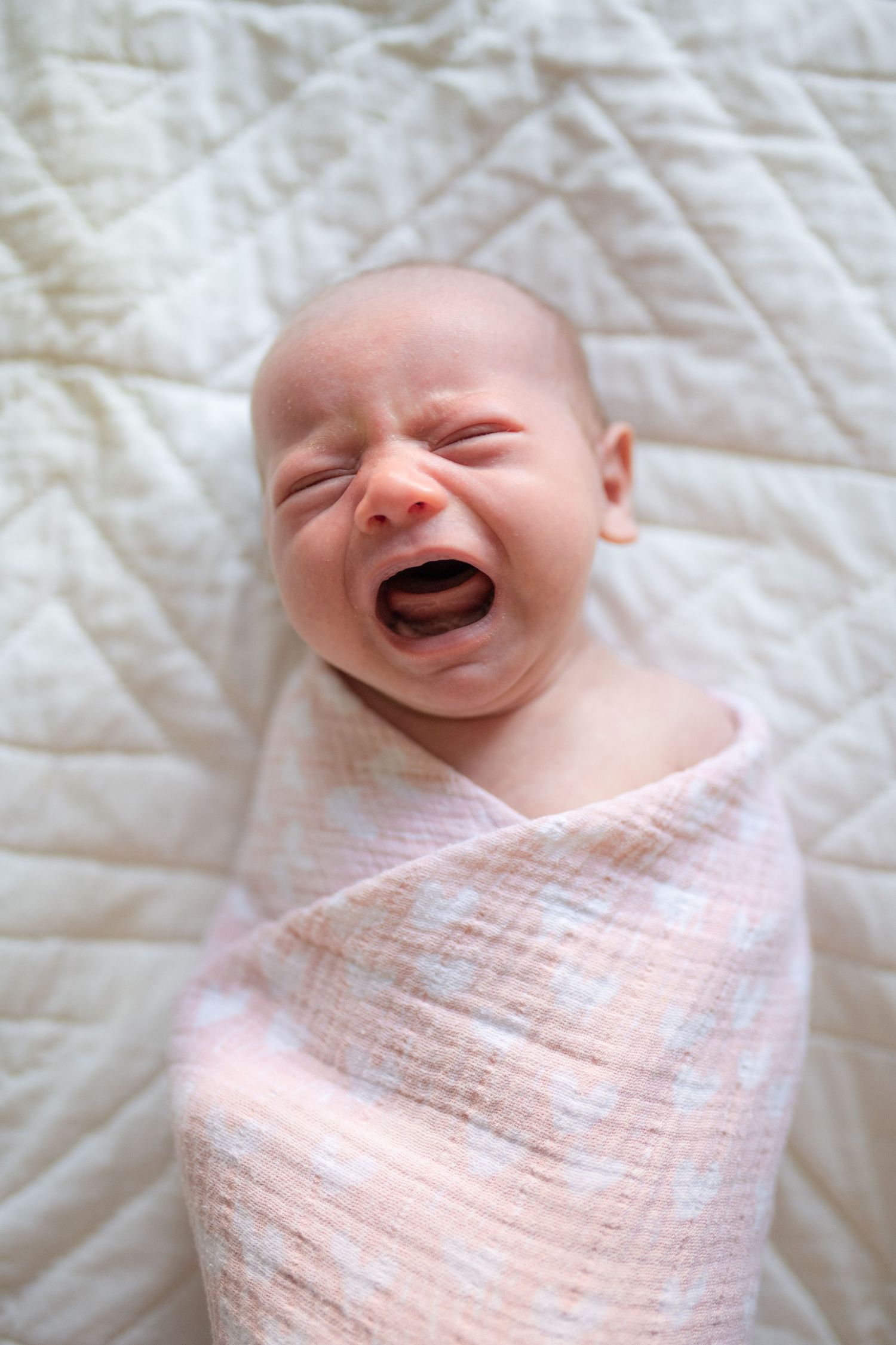 A crying newborn baby swaddled in a soft pink blanket lies on a white quilted surface.