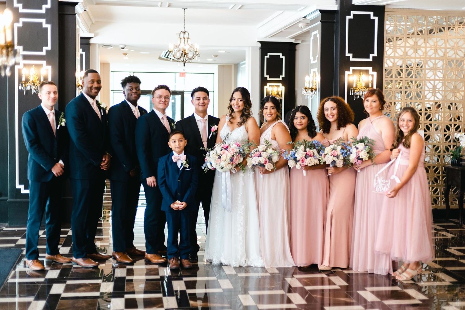 Wedding party in navy suits and blush pink bridesmaid dresses poses together in an elegant venue with checkered floors.