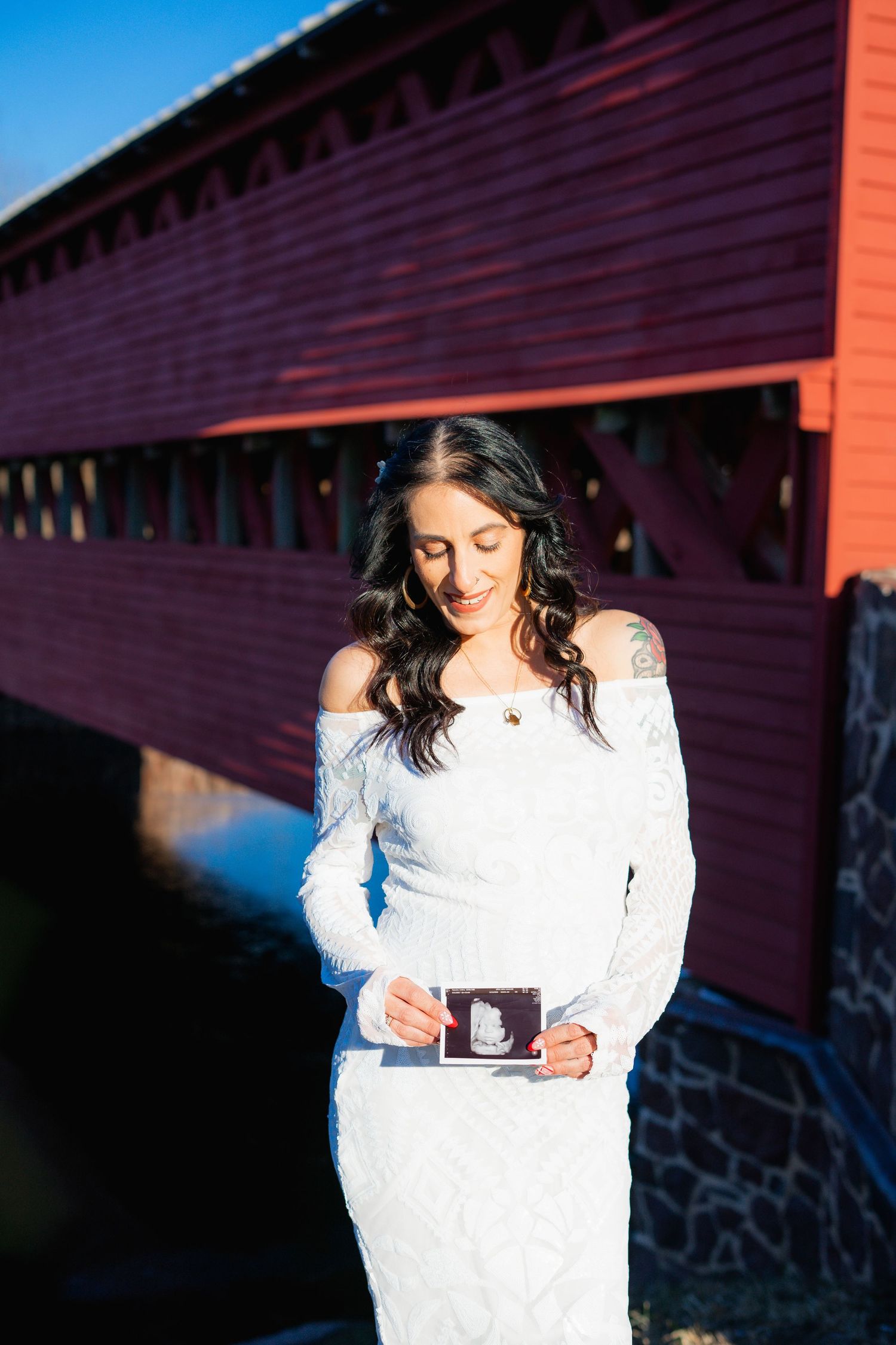 Woman in white lace dress holds ultrasound photo while standing in front of red covered bridge in sunlight.