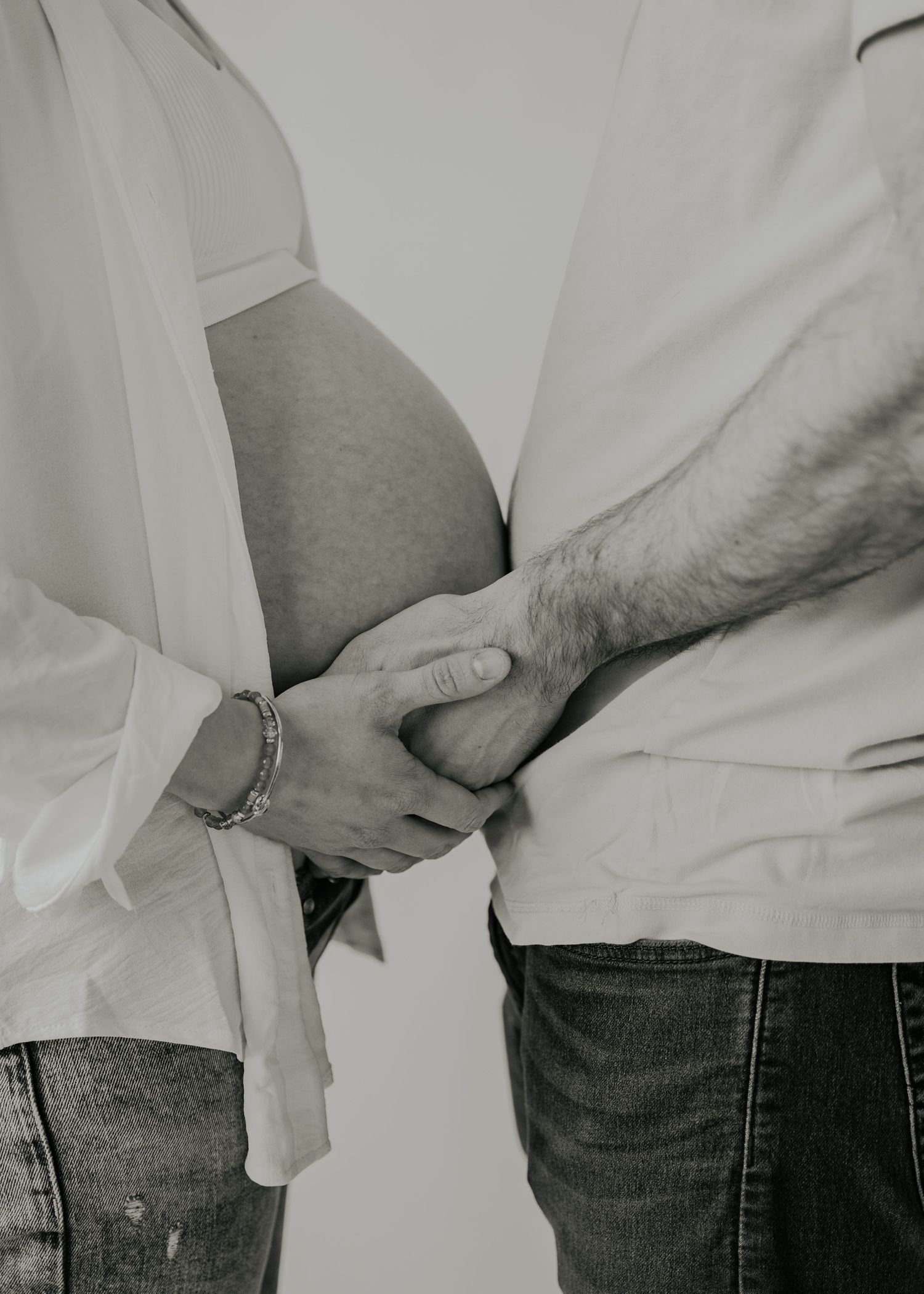 Close-up photo of expecting parents holding hands over pregnant belly wearing white shirts and jeans.