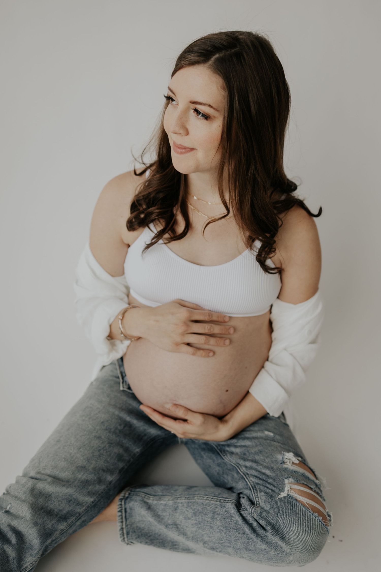 Expecting woman in white cropped top and ripped jeans gently holds her pregnant belly while sitting against neutral wall.