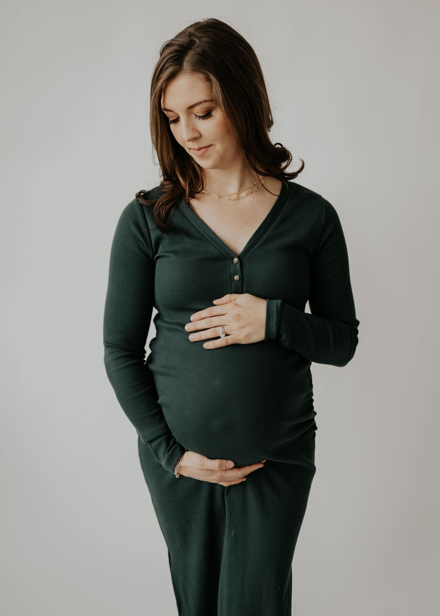 Expectant mother wearing dark green maternity sweater and pants poses against neutral backdrop while holding her belly.