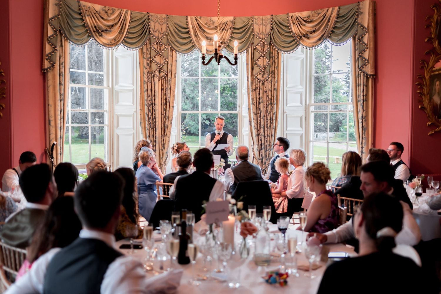 Wedding guests seated at an elegant dinner reception in a grand room with pink walls and traditional draped windows.