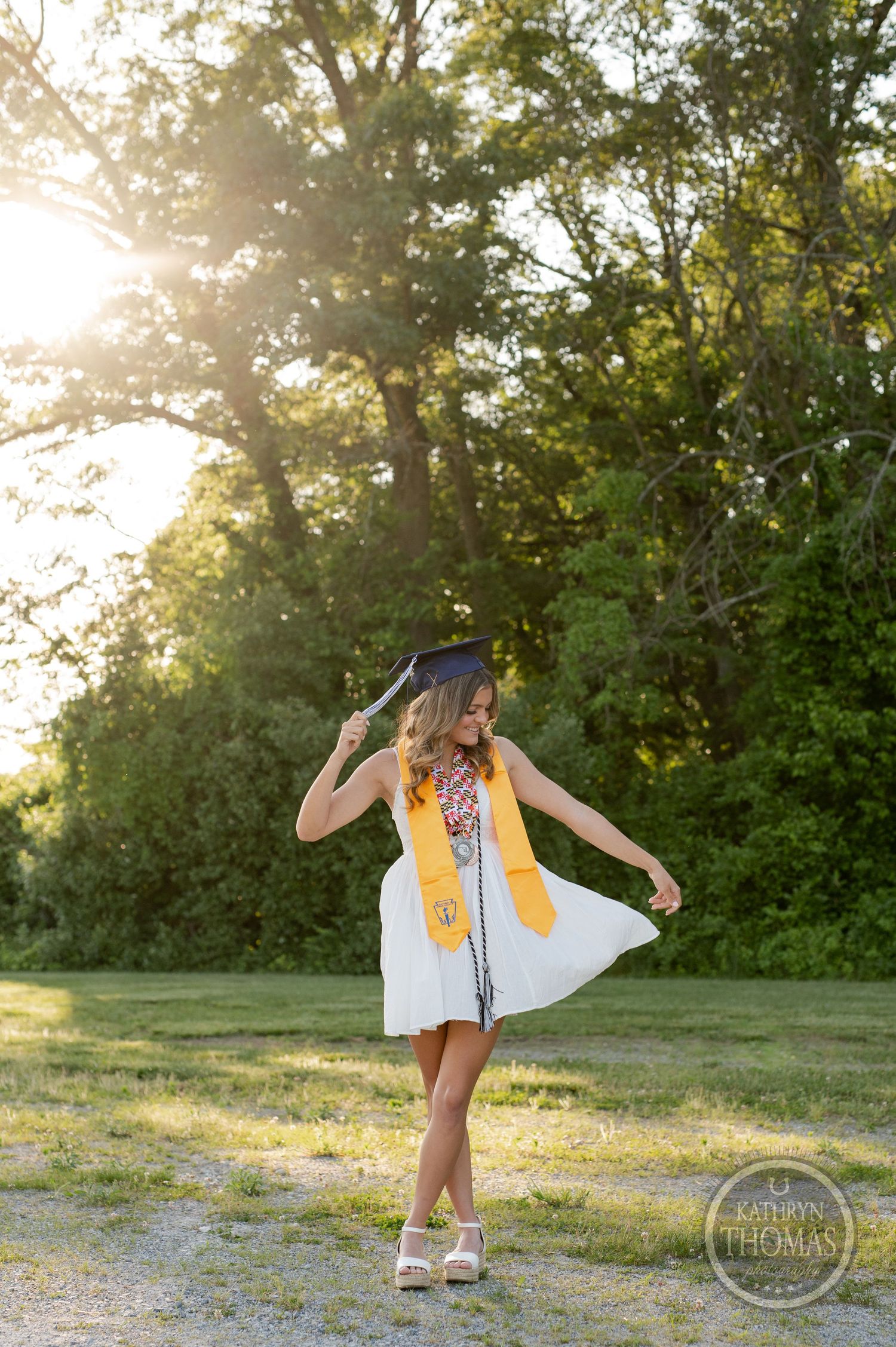 Graduate in white dress and academic regalia twirls playfully in sunlit outdoor setting.
