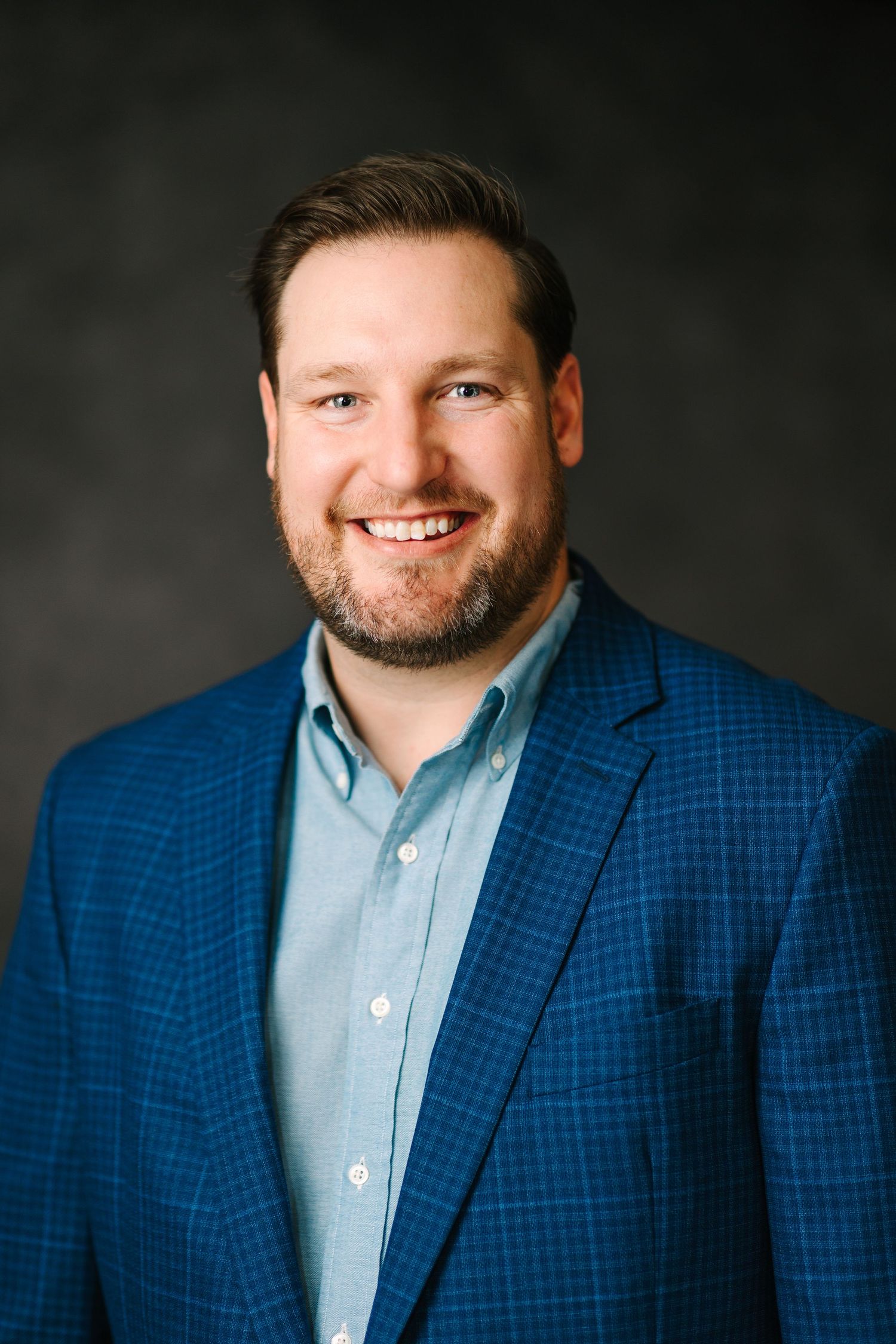 Professional headshot of a person wearing a blue checkered blazer against a dark background.