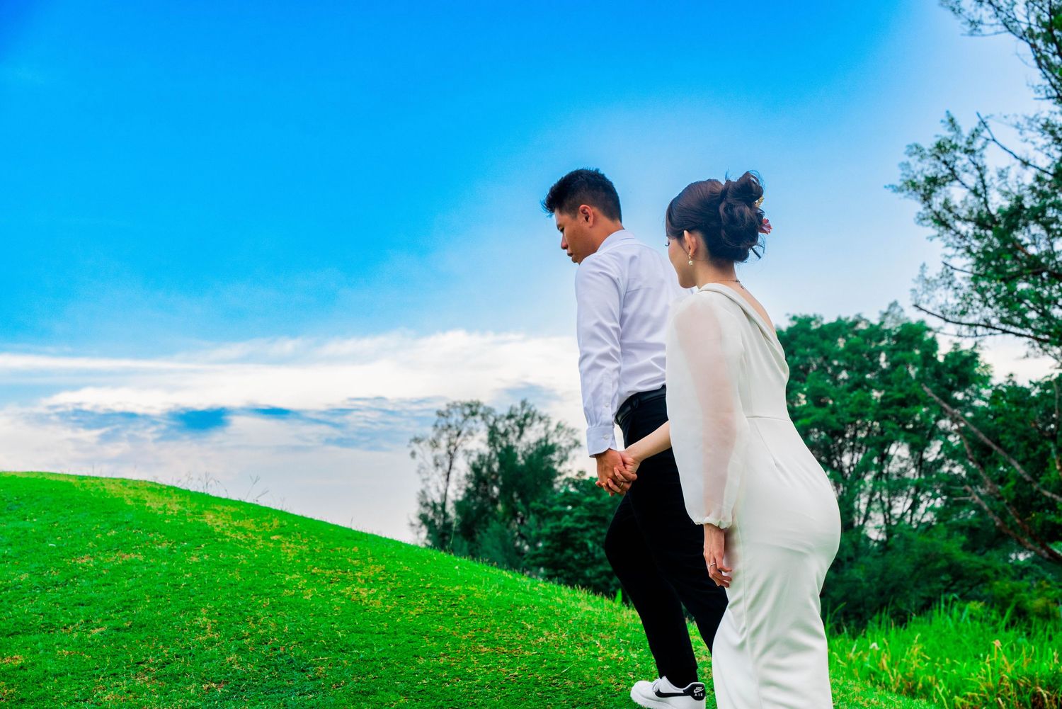 Couple in wedding attire walking on green hillside against blue sky.