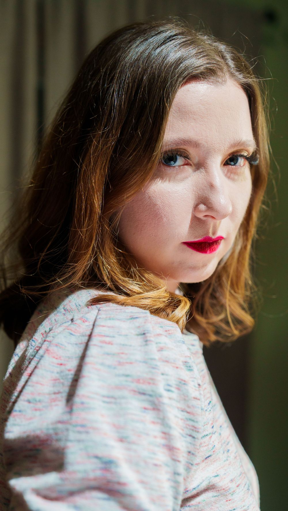 Portrait of someone wearing red lipstick and a textured white sweater, looking over their shoulder in soft natural lighting.