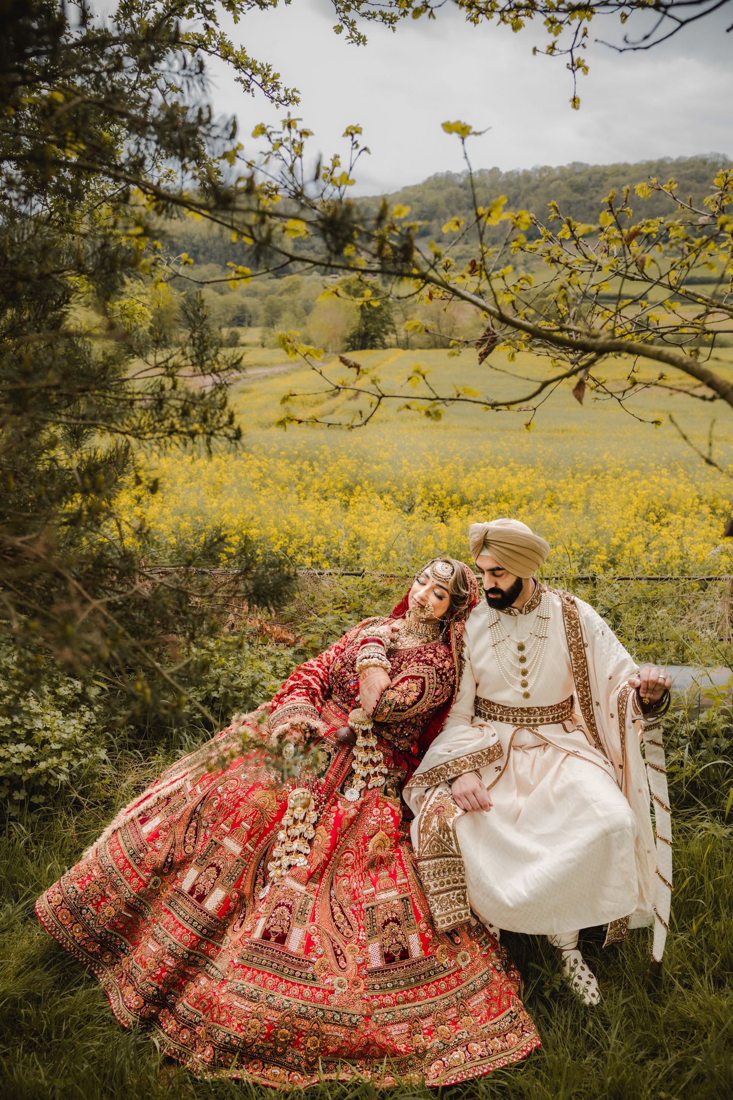 A couple in traditional Indian wedding attire sits together in a meadow with yellow wildflowers and rolling hills.