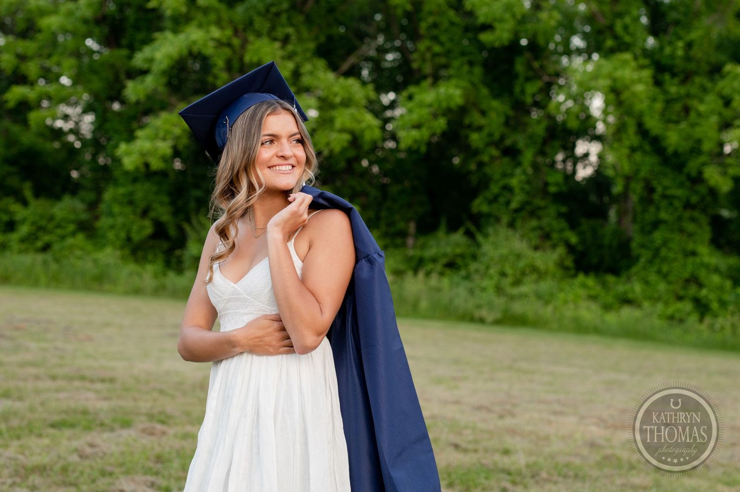Graduate in white dress and navy cap poses thoughtfully against green background.