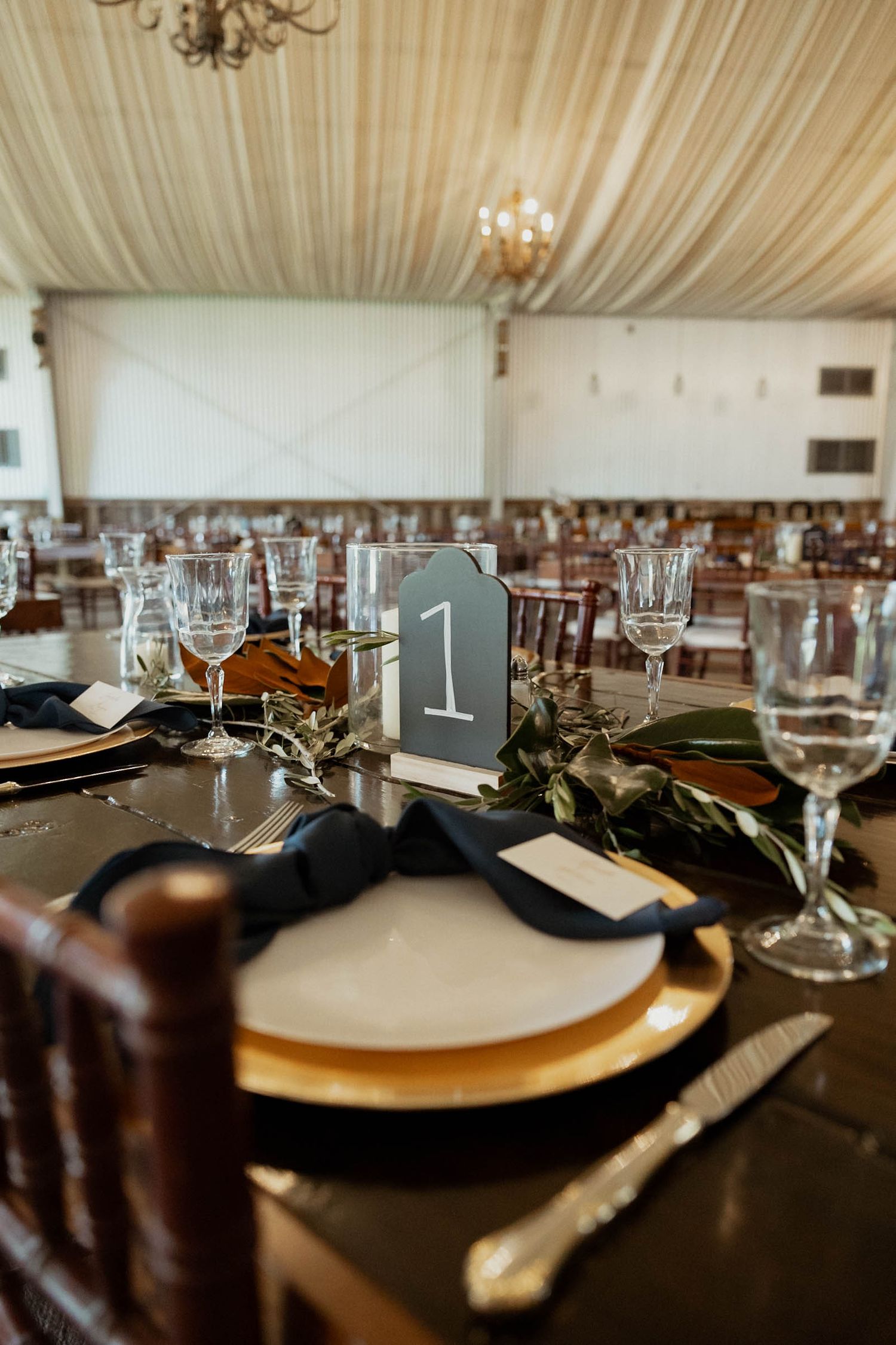 Elegant wedding reception table setting with gold chargers, black napkins, and greenery runner.