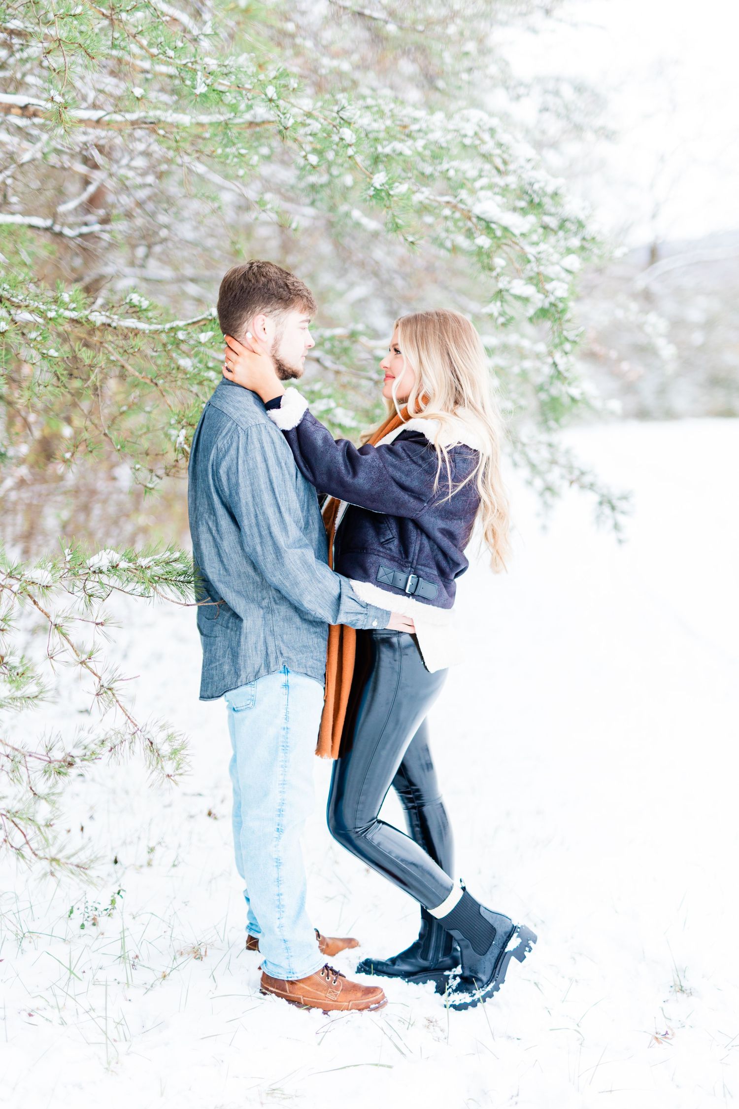 A couple shares a tender moment while standing in fresh snow surrounded by evergreen branches.