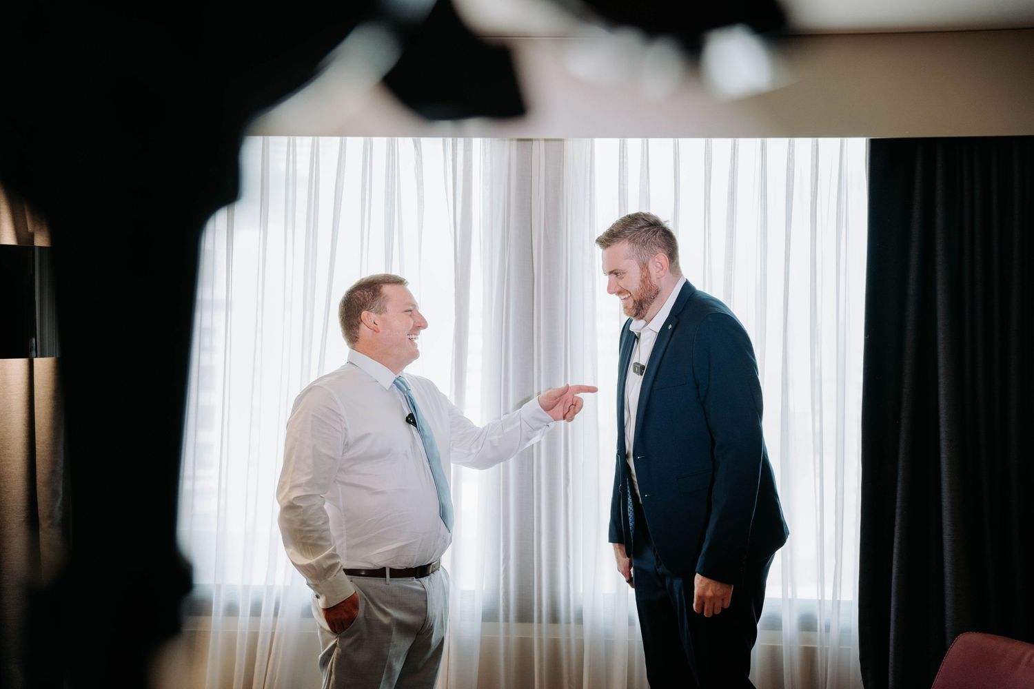 Two men in formal attire having an animated conversation in front of white curtains.