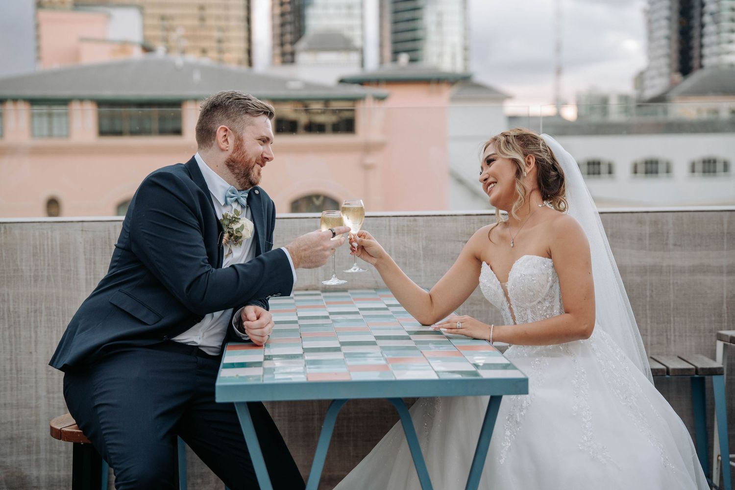 A wedding couple shares drinks at a rooftop table in an urban setting.