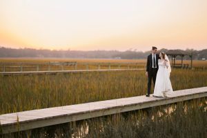 A couple in wedding attire stands on a wooden boardwalk through marsh grass at sunset.