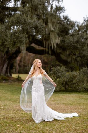 A bride in a lace mermaid wedding dress and veil poses gracefully in a garden with Spanish moss draped oak trees.