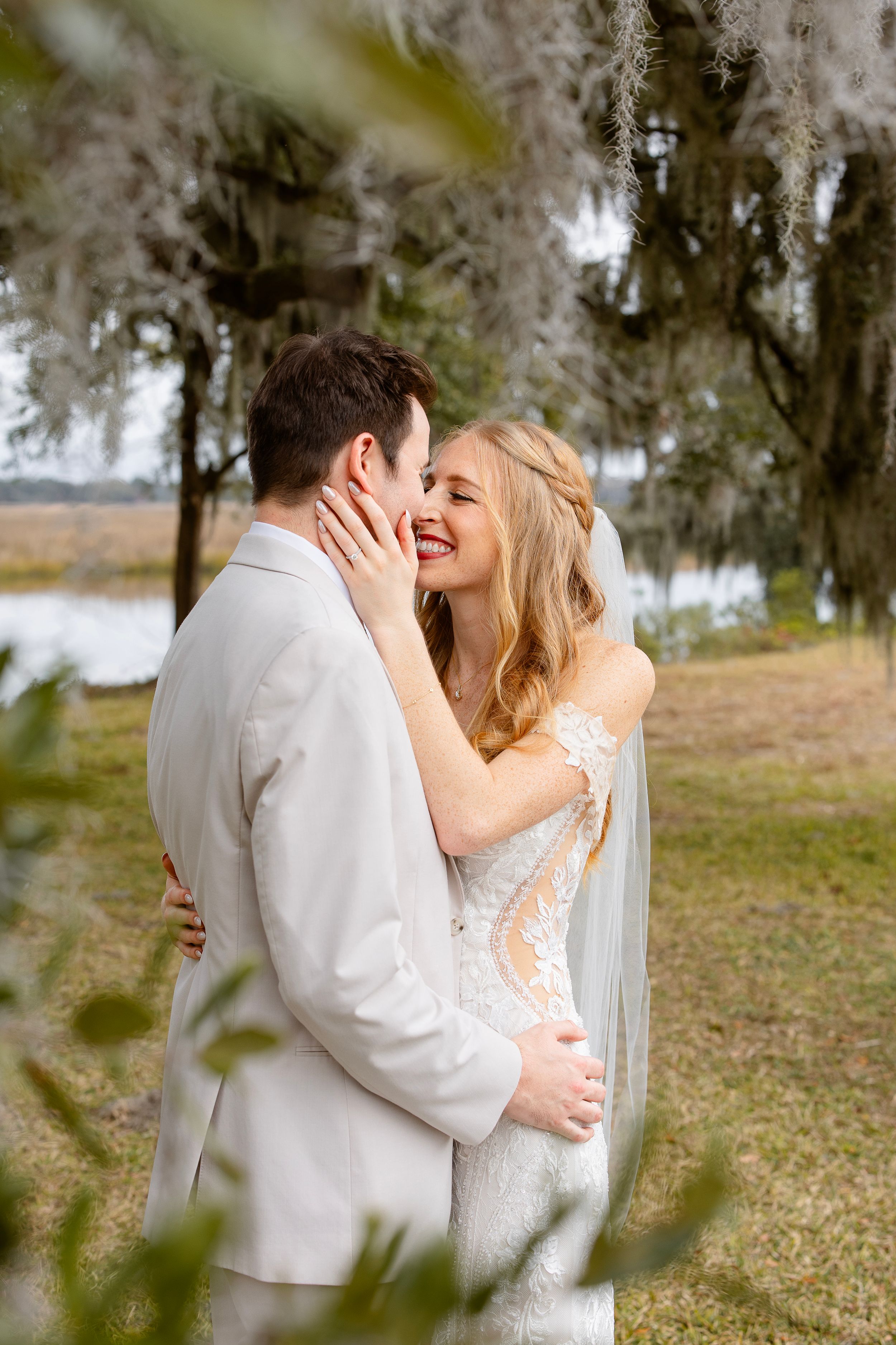 A romantic moment between newlyweds embracing outdoors under Spanish moss-draped trees near a waterfront.