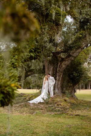 A wedding couple stands together beneath a majestic moss-draped oak tree in a serene outdoor setting.