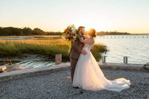 Couple embraces at sunset by a waterfront dock with golden light streaming through marsh grass.