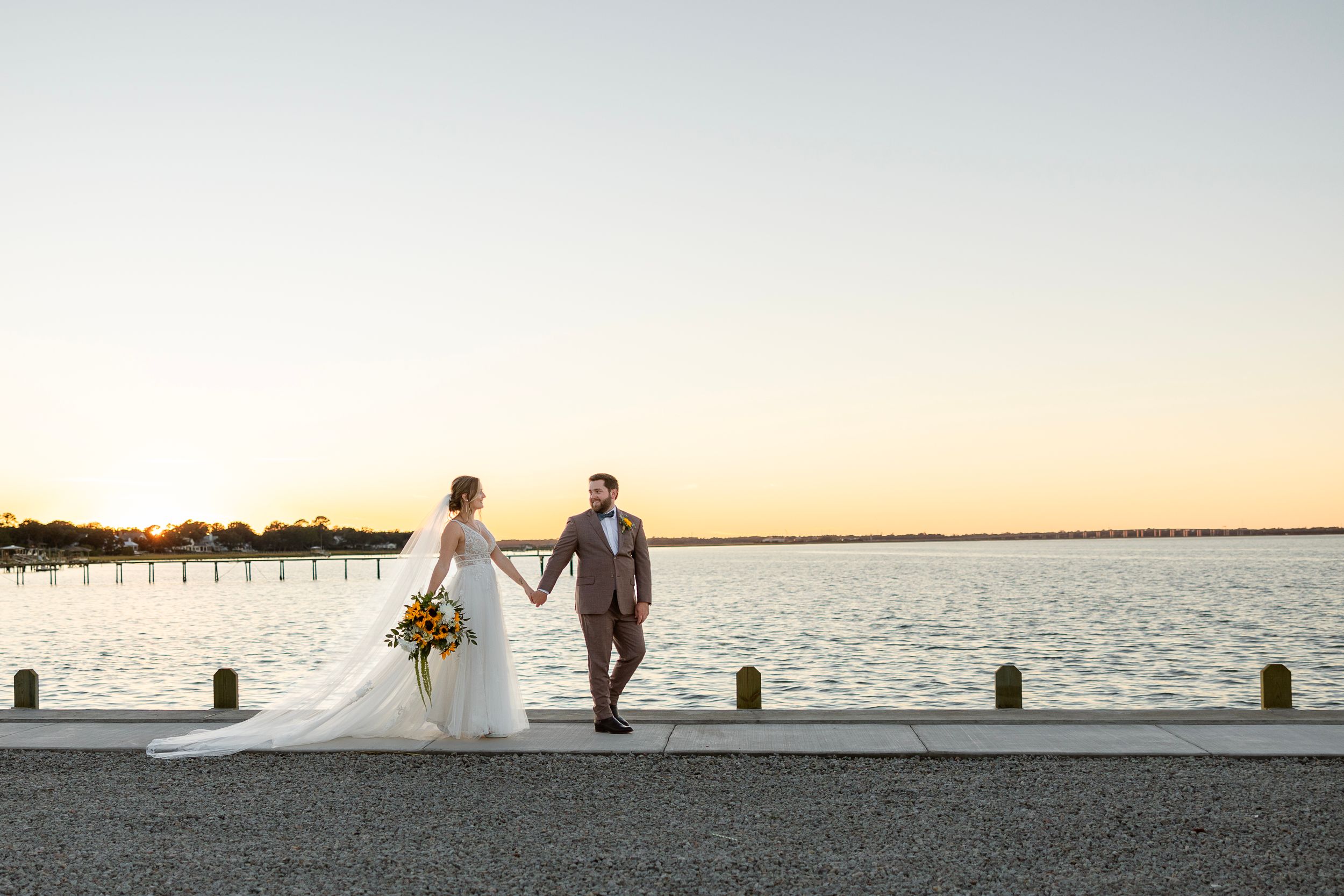 A romantic sunset waterfront scene with a couple in wedding attire walking along a pier at dusk.